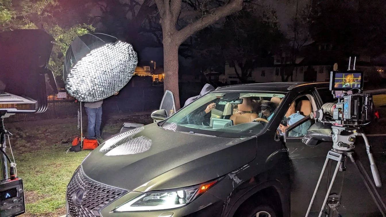 Night exterior shoot with a dark SUV, overhead diffusion rig, camera on tripod, crew, and lit residential windows