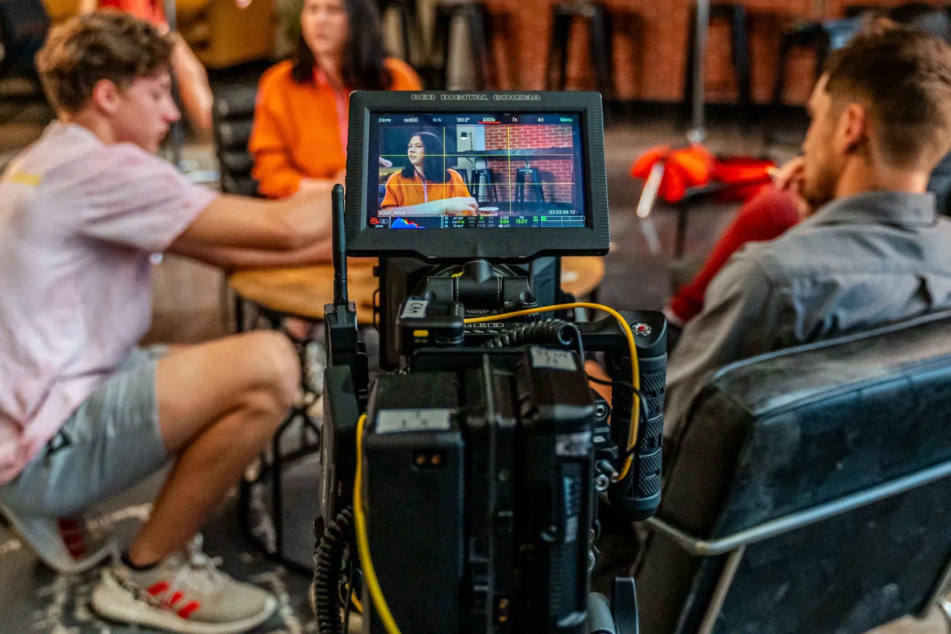 Viewfinder view from a RED cinema camera of a woman in an orange top seated at a table against a brick wall
